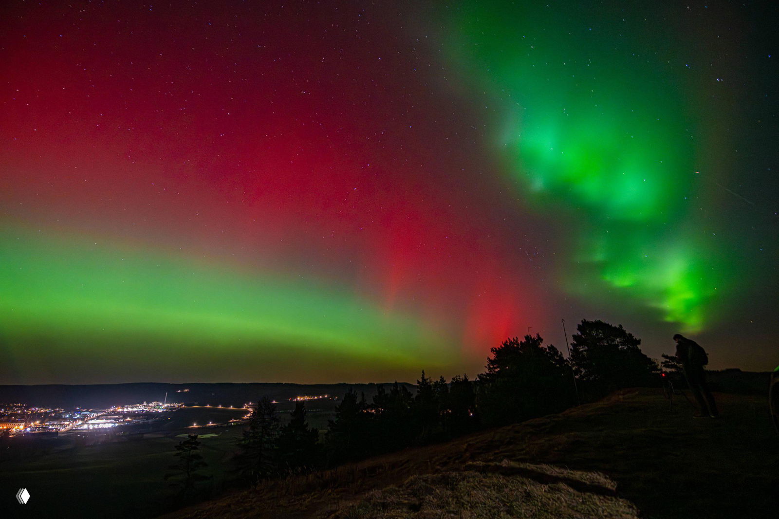 Многоцветные ленты северного сияния с зелёными и красными полосами над холмистой местностью; справа виден силуэт фотографа на краю съёмки.