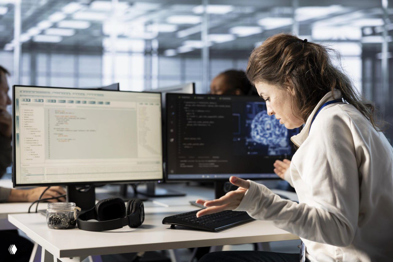 Женщина-специалист за столом в офисе перед двумя мониторами: на экранах код и графика нейросети, рабочая сцена.