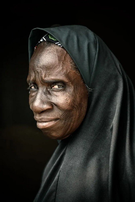 Крупный художественный портрет пожилой женщины в тёмном платке: глубокие морщины, выразительный взгляд и фактура кожи на нейтральном фоне.