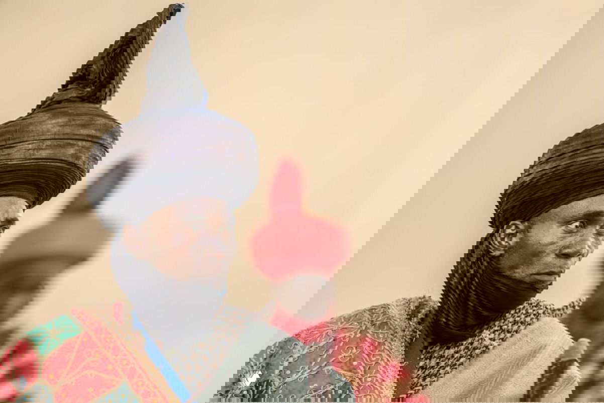 Портрет мужчины в высоком турбане и яркой традиционной одежде, на размытом фоне виден второй человек; акцент на текстурах и цвете костюма.