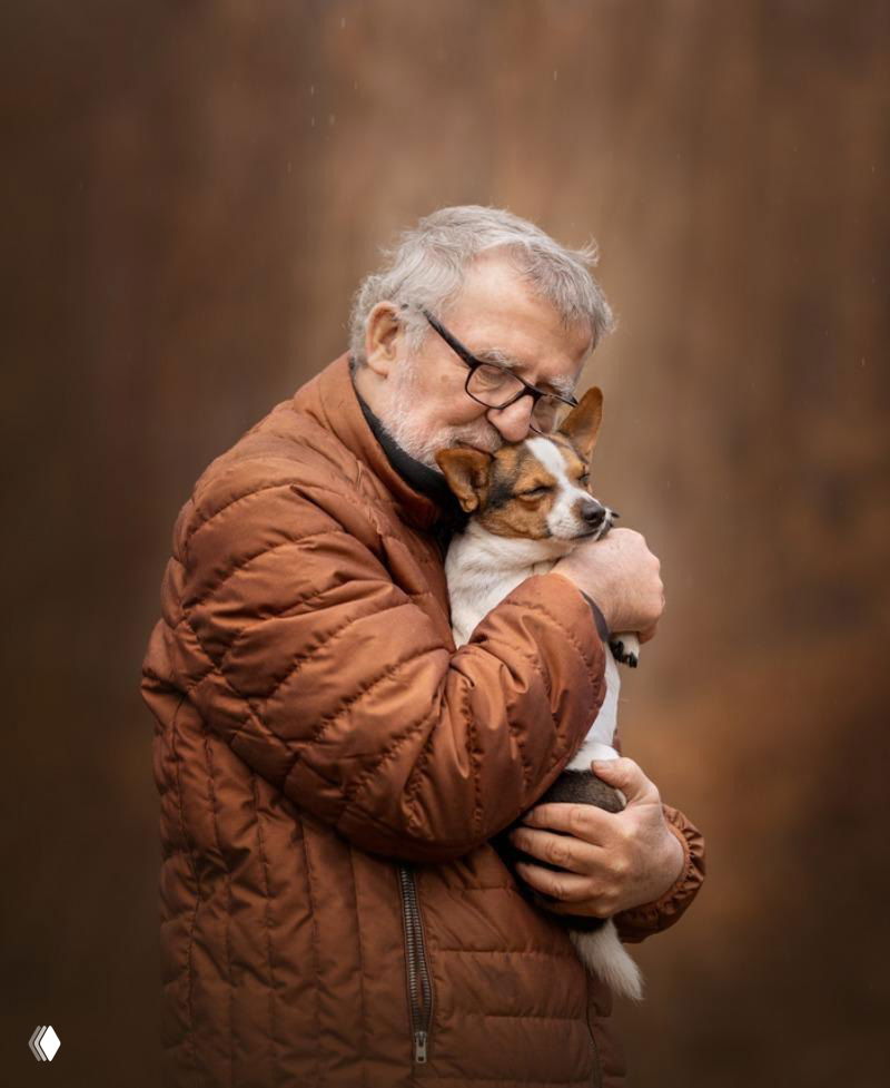 Портрет пожилого мужчины в коричневой стёганой куртке, нежно прижимающего небольшую собаку к груди на тёплом размытом фоне.