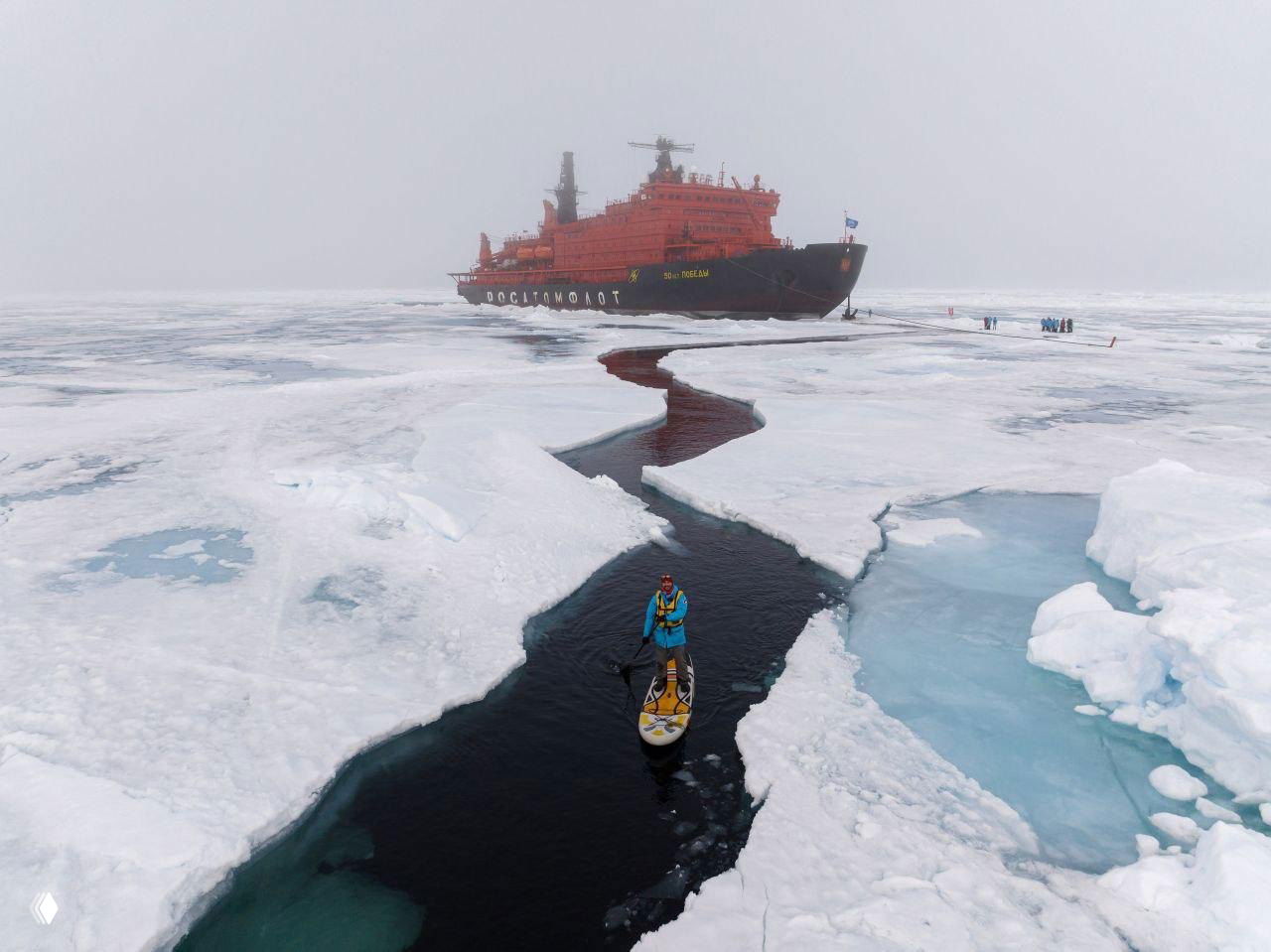 Атомный ледокол оранжевого цвета среди арктических льдин; на переднем плане человек на сапборде в узком проливе.