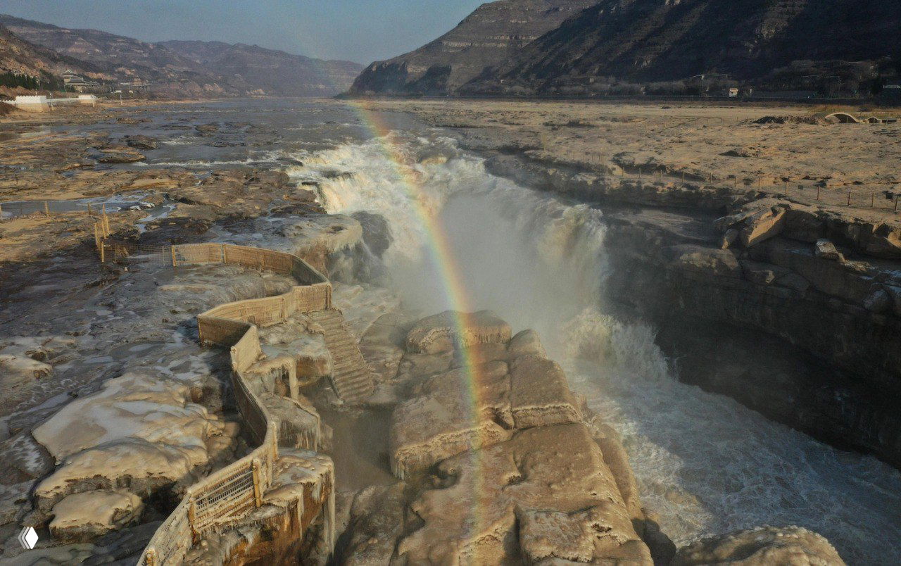 Вид сверху на водопад Хукоу и яркую радугу: замёрзшие террасы, бурная река Хуанхэ и дуга света на фоне зимнего ландшафта.