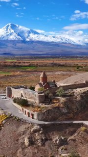 Аэрофотоснимок монастыря Хор Вирап на скале с подъездной дорогой и крепостными стенами, на фоне большой заснеженной горы под голубым небом.