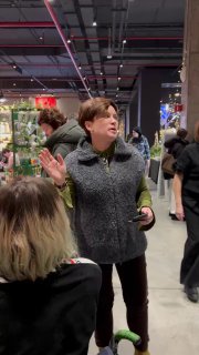 Организатор у прилавка на GreenMarket беседует с посетителями; в кадре видно праздничное оформление, торговые столы с кашпо и декор.