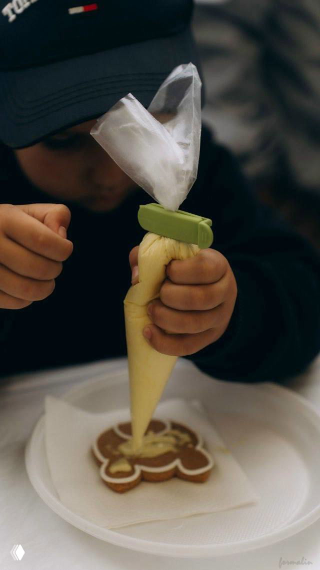 Детская рука с кондитерским мешком наносит белую глазурь на пряник в форме человечка во время мастер‑класса по росписи пряников.