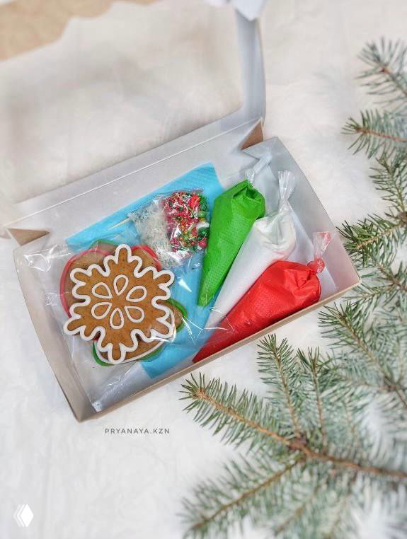 Подарочная коробка с украшенным пряником и тюбиками цветной глазури в праздничной упаковке рядом с еловой веткой.