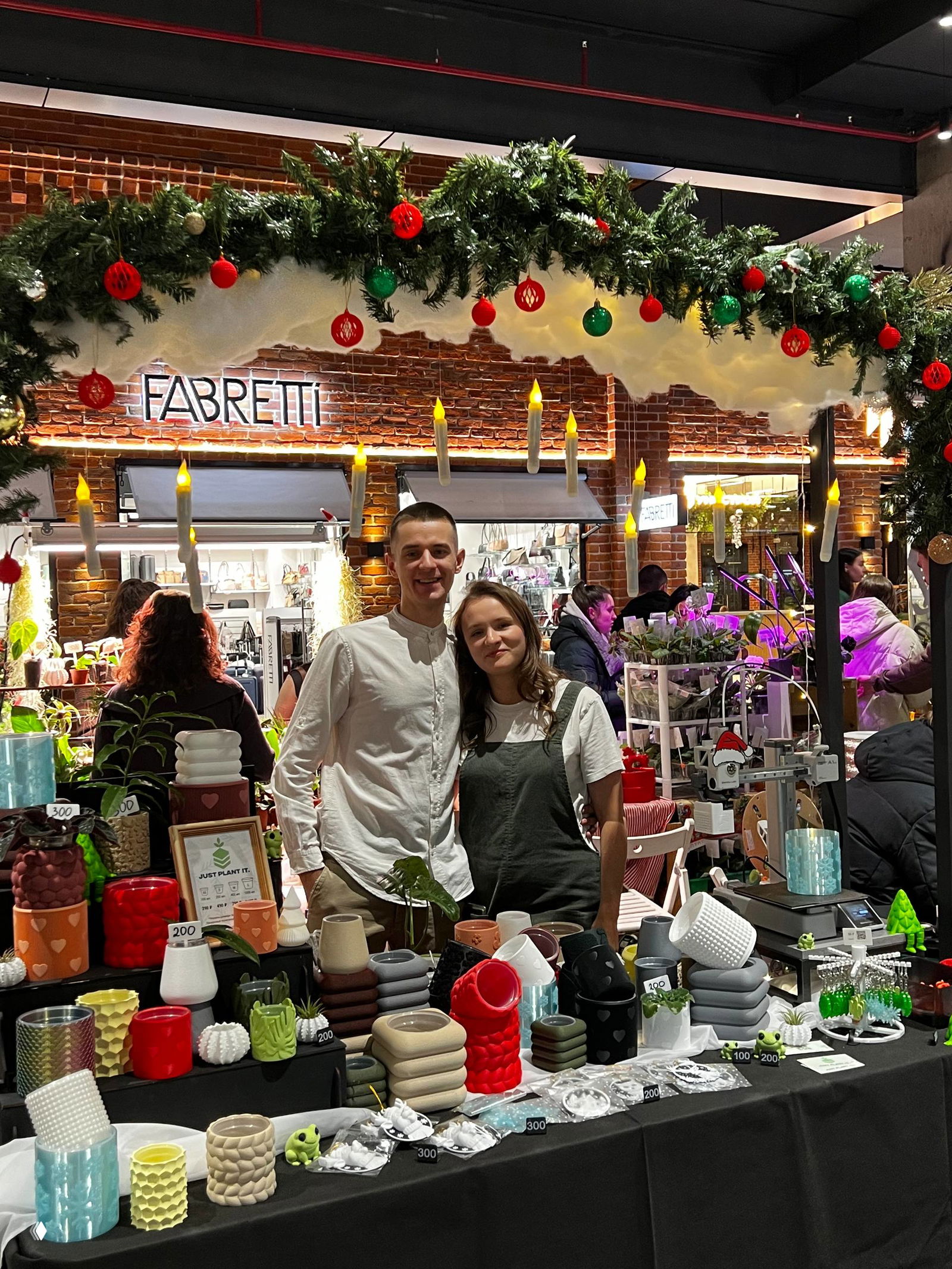 Пара у прилавка «Just plant it» на фоне новогодней гирлянды; ассортимент кашпо, аксессуаров и небольшой 3D‑принтер на столе.