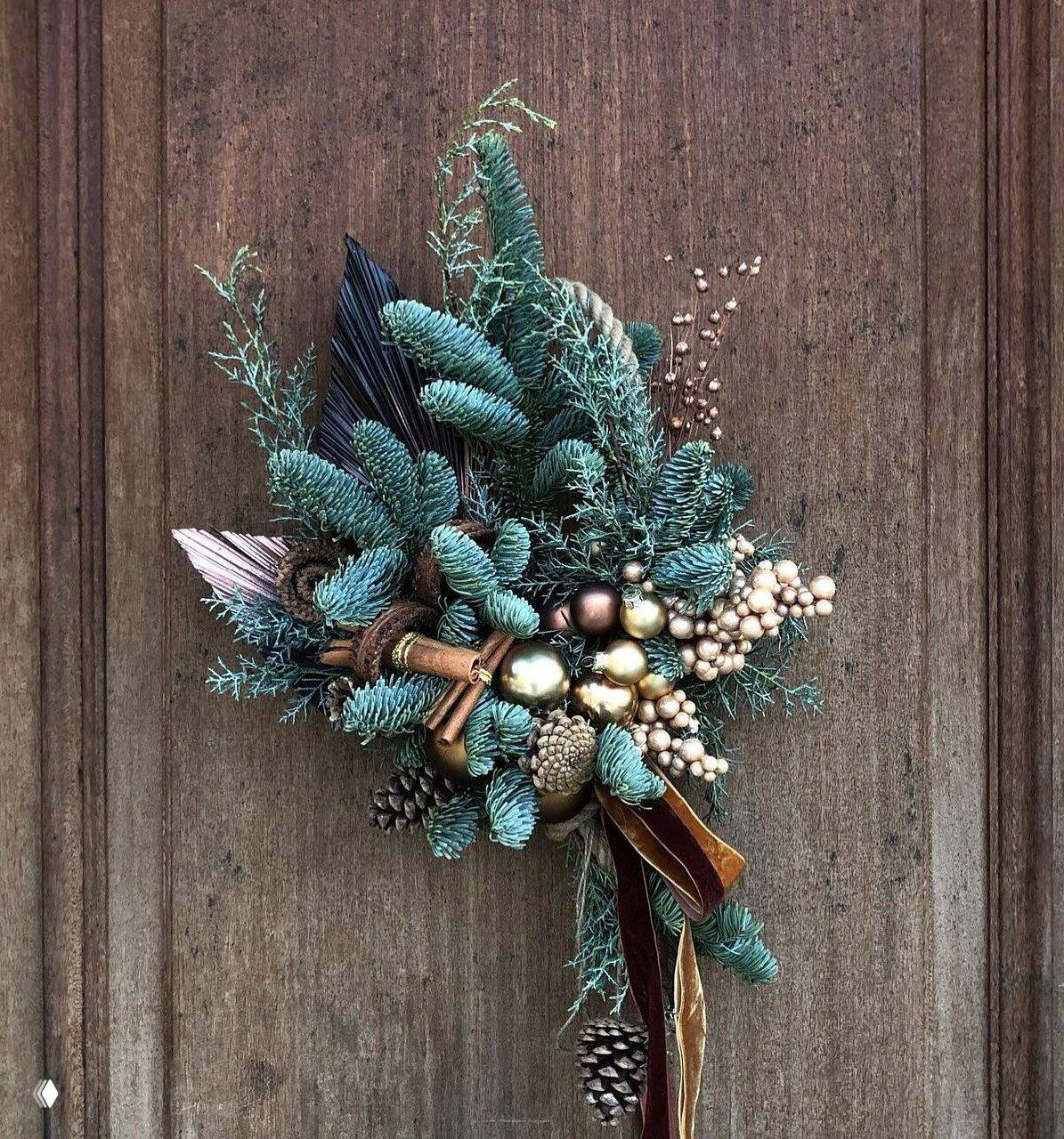 Новогодняя подвеска из еловых веток, шишек, золотых шариков и бархатных лент на деревянной двери, дополненная корицей и декоративными ягодами