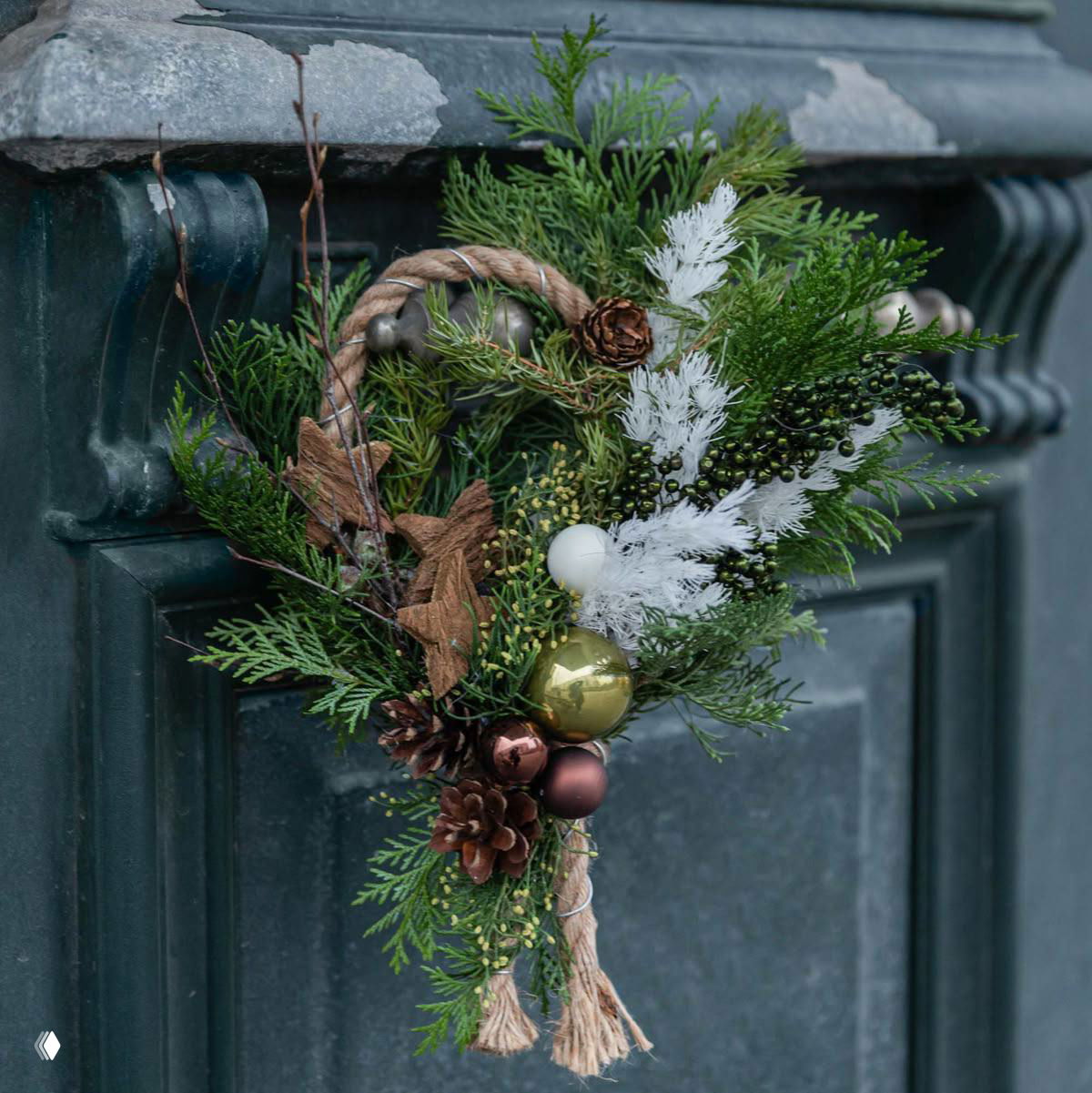 Праздничная дверная петля из хвойных веток и каната с кедровыми шишками, белыми перьевидными акцентами и золотыми шариками на металлическом фоне