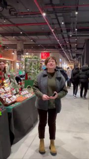 Посетитель стоит у прилавков на цветочном маркете в закрытом помещении: видно торговые столы с растениями, декором и нескольких прохожих на фоне.