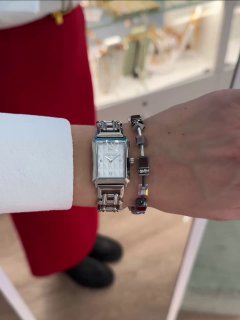 Запястье в бутике: прямоугольные серебристые часы с металлическим браслетом в сочетании с тонким декоративным браслетом на фоне витрины.