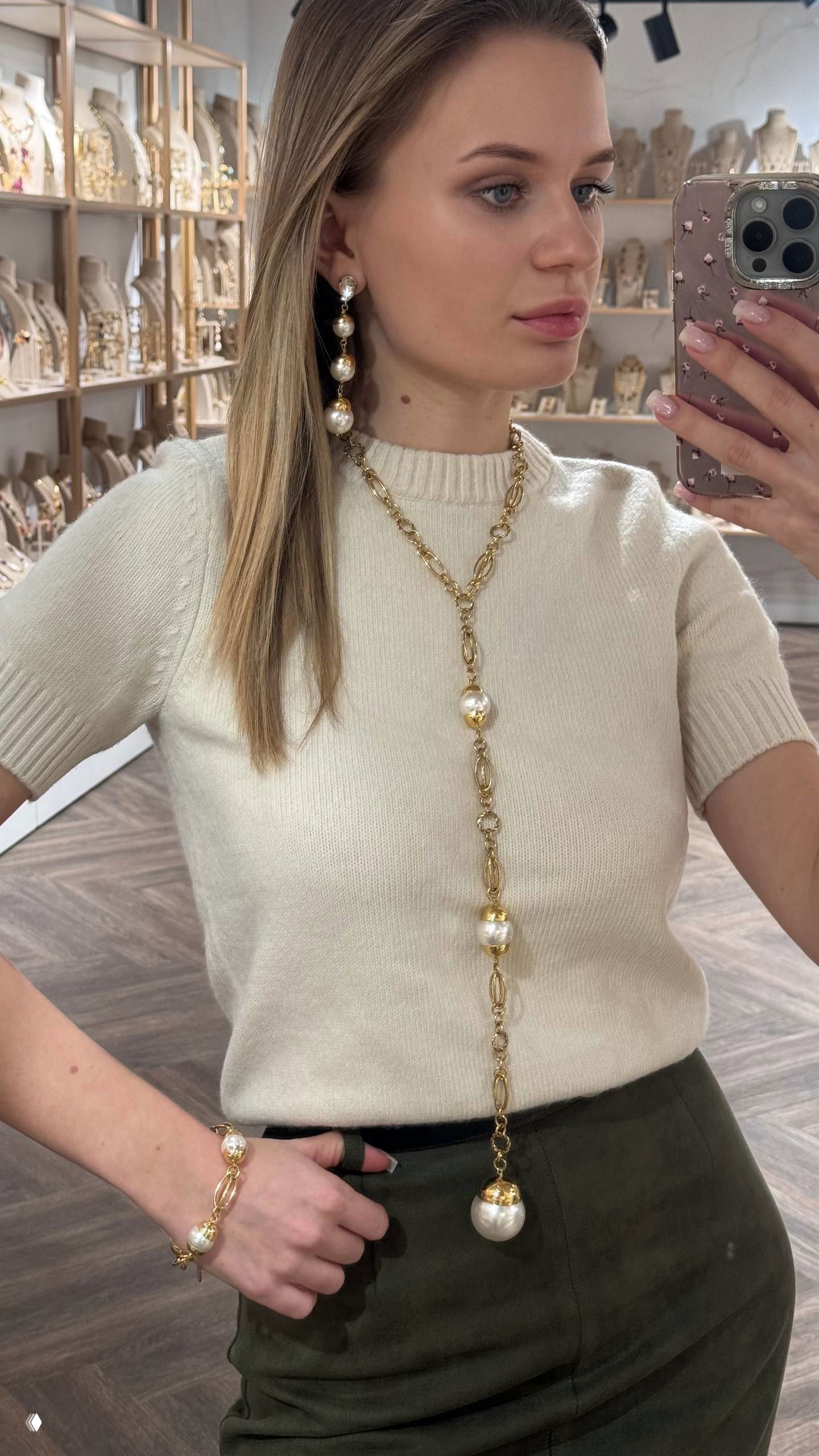 Модель в светлом свитере демонстрирует длинное золотое колье с подвесами и браслет, снимок сделан в бутике Rosone у зеркала.
