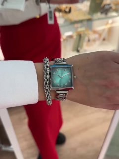 Запястье на фоне продавца в красной форме: квадратные часы с зелёным градиентным циферблатом и массивным браслетом рядом с цепочкой.