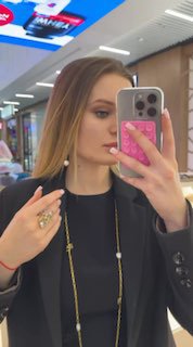 Селфи в зеркале в торговом центре: девушка в черном пиджаке носит длинную золотую цепь с жемчужным кулоном и демонстрирует кольцо на пальце.