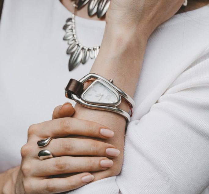Женское запястье в серебристых часах UNOde50 и комплектных украшениях на светлом фоне — стилизованная съёмка аксессуаров и гардероба.