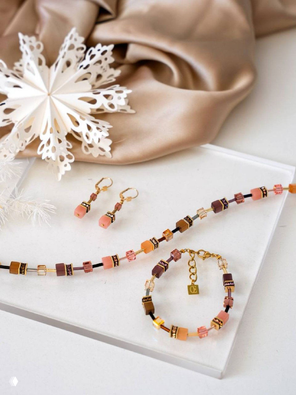 Нежная композиция: бусы, браслет и серьги Coeur de Lion на светлом подносе рядом с декоративной бумажной снежинкой.