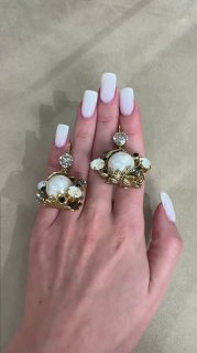 Крупный план руки с белым маникюром, демонстрирующей массивные золотые кольца с крупными круглым жемчугом и декоративными элементами.