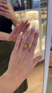 Крупный план руки с аккуратным маникюром и массивным золотым кольцом Claudio Canzian; отражение в зеркале и прилавок с украшениями видны позади.