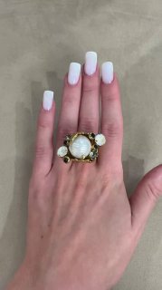 Рука с аккуратным белым маникюром и крупным золотым кольцом с барочной жемчужиной, фон нейтральный, акцент на форме и текстуре жемчуга.