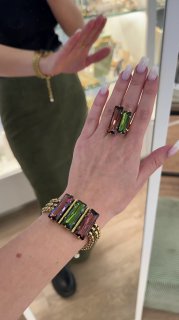 Крупный план руки с изумрудно-зелёным крупным камнем в кольце и браслетом Claudio Canzian, на заднем плане витрина с подсвеченными ювелирными изделиями.