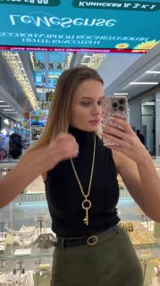 Женщина у зеркала демонстрирует длинную цепь с подвеской-ключом и серьги Claudio Canzian в торговом центре, видны торговые ряды и вывески сверху.
