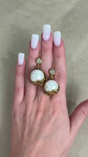 Рука с белым маникюром удерживает пару массивных золотых серег с круглыми жемчужинами — снимок сделан на нейтральном фоне для акцента на украшении.