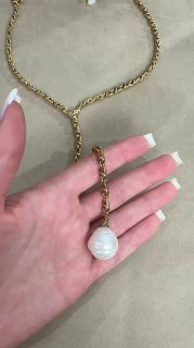 Рука держит золотую цепочку с крупным белым жемчужным подвесом на фоне бежевого бархата — акцент на форме кулона и звеньях цепи.