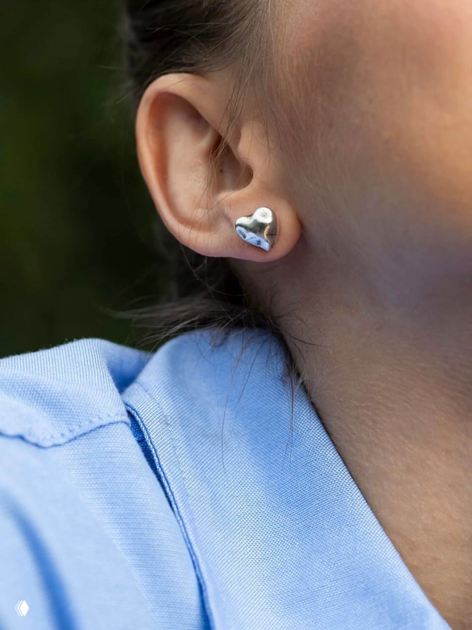 Макро‑съёмка сережки‑пуссета в виде выпуклого серебряного сердца на мочке уха, мягкий боке на заднем плане.