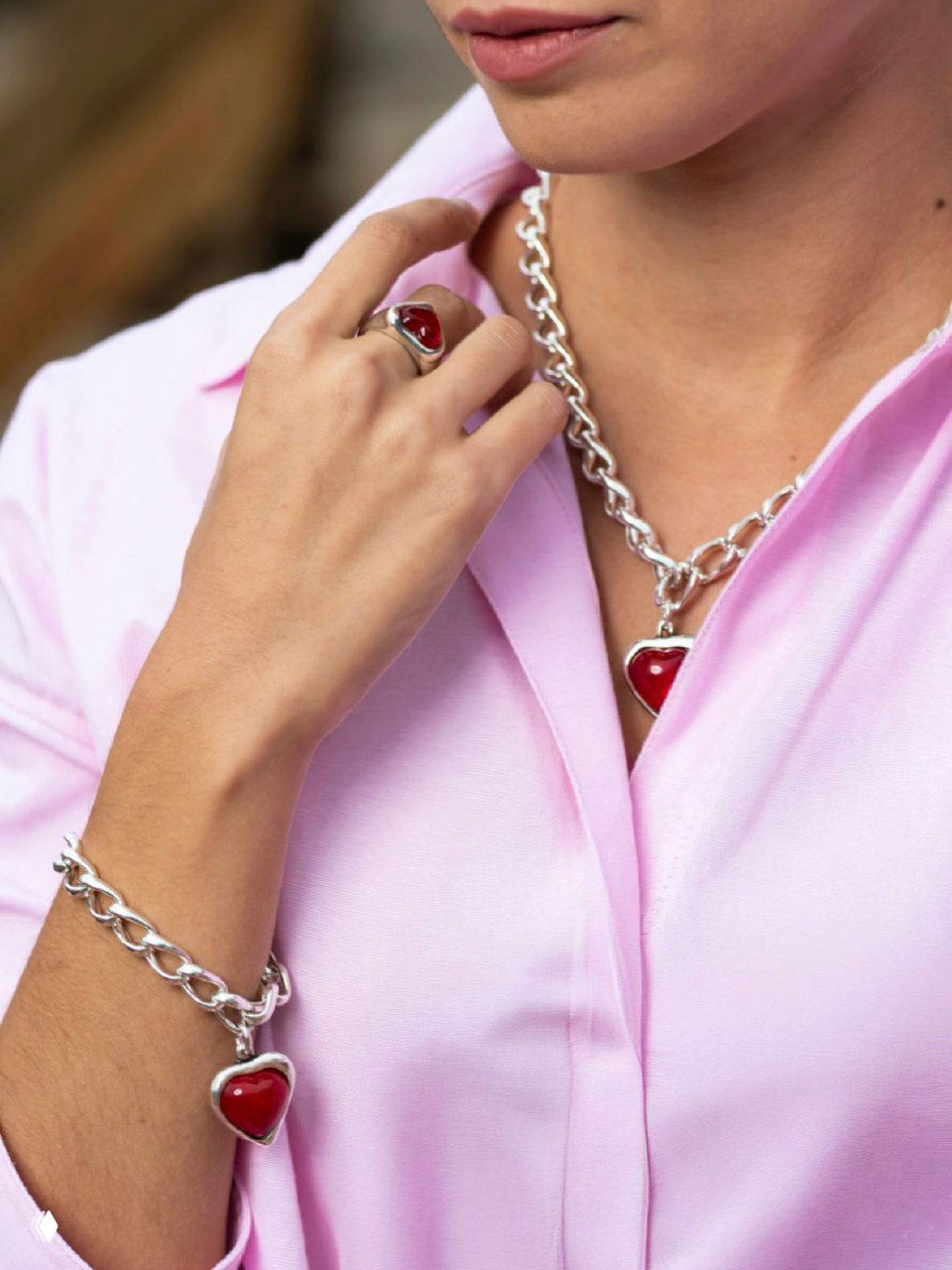 Женщина в розовой блузке демонстрирует массивное серебряное колье с ярко‑красной сердцевидной подвеской и подходящее кольцо на пальце.
