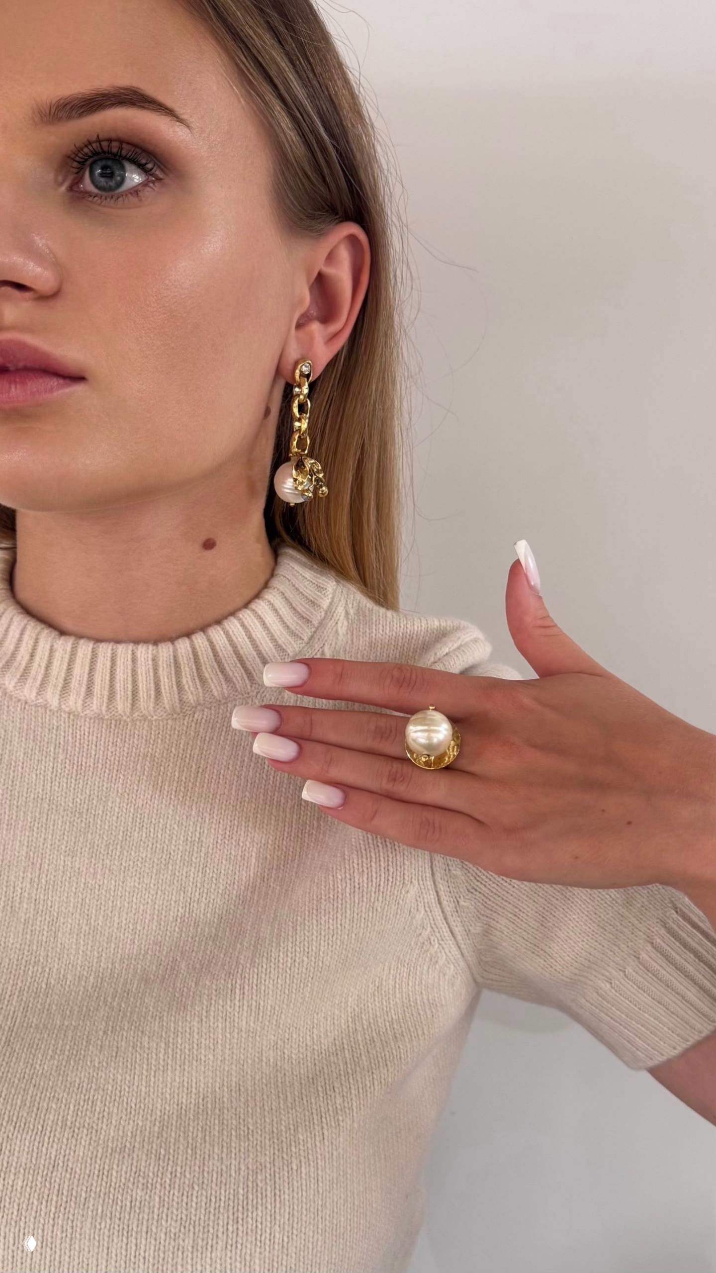 Портрет девушки в светлом свитере, на ухе свисающая золотая серьга с большим жемчужным кулоном и рука с крупным кольцом на фоне нейтральной стены.