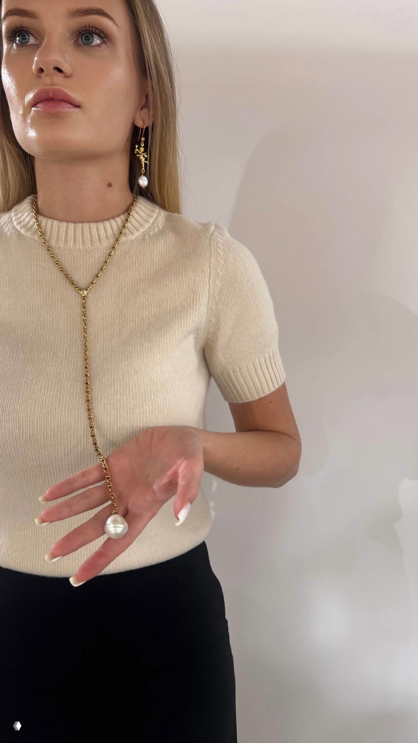Девушка в светлом свитере держит в руке длинное золотое колье с одним крупным жемчужным подвесом — студийная съемка с мягким светом.