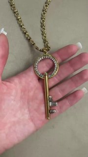 Крупный план женской руки с аккуратным маникюром держит золотистую подвеску-ключ с инкрустацией кристаллами на нейтральном фоне.