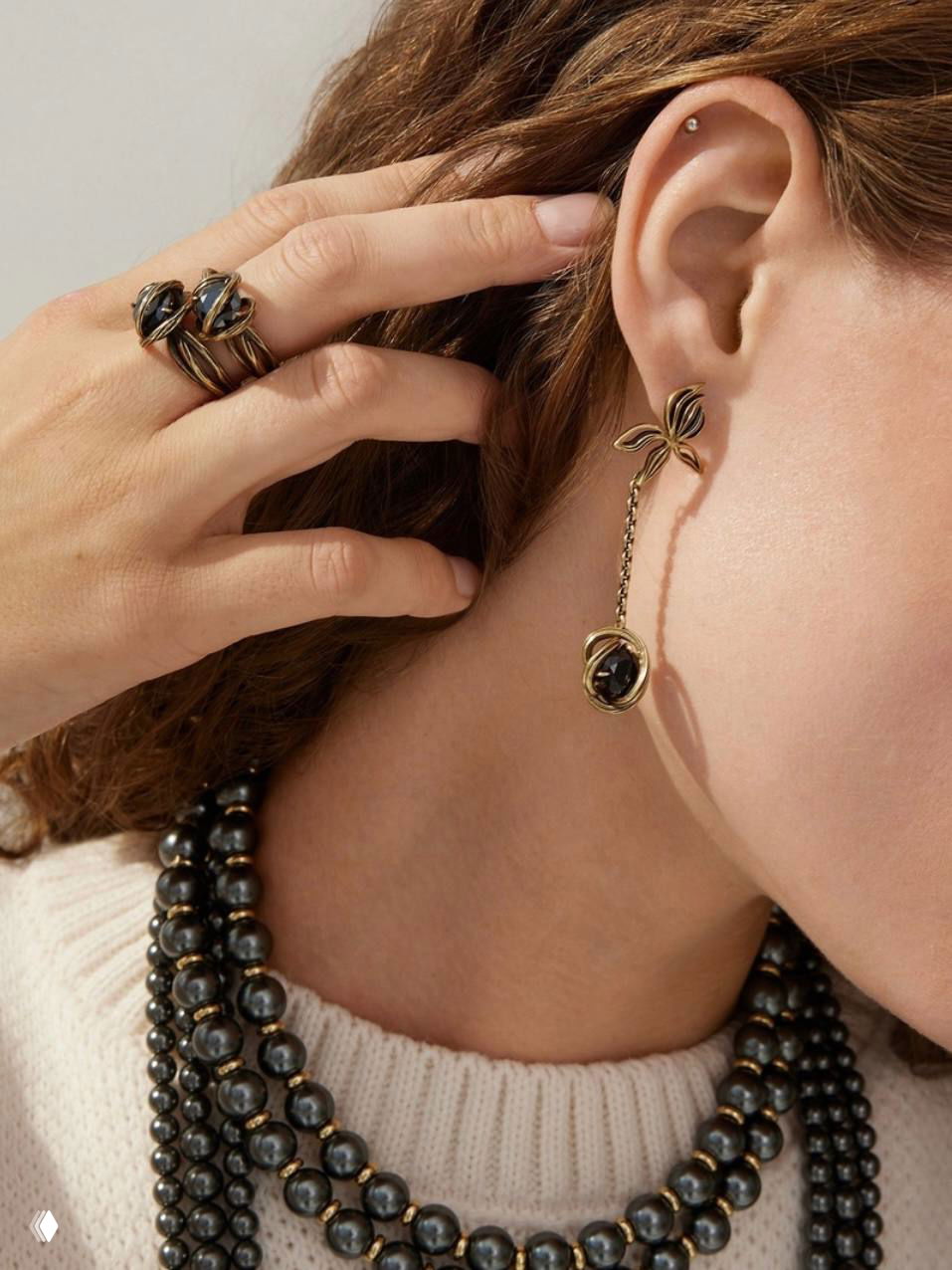 Боковой крупный план уха и шеи модели с длинной подвесной серьгой, многорядным тёмным жемчужным ожерельем и кольцами — детали коллекции Fiore di Firenze.