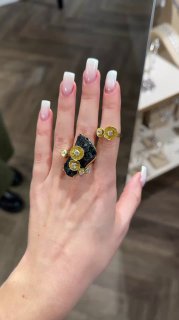 Рука с французским маникюром держит и показывает комплект ювелирных изделий KALLIOPE: кольца и декоративные элементы на черном фоне магазина.