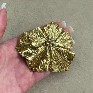 Крупный план золотой броши в форме цветка в ладони: рельефная текстура лепестков, блеск и художественная обработка металла.