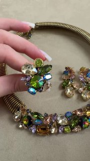 Рука держит комплект Claudio Canzian: колье и серьги на бежевом фоне, видны зелёные и разноцветные кристаллы и металлическая основа украшений