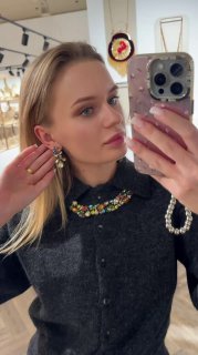 Девушка в бутике делает селфи и демонстрирует украшения Claudio Canzian: колье и серьги с зелёными и разноцветными кристаллами, витрина на заднем плане