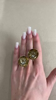 Крупный план двух золотистых круглых серёг, держимых на руке с белым маникюром; видно глянцевую поверхность, крепления и внутреннюю фактуру.