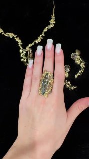 Рука с большим декоративным кольцом демонстрирует украшения на чёрном фоне; видна инкрустация и форма кольца, фокус на текстуре металла.