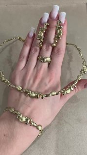 Рука с маникюром держит комплект из браслетов и ожерелий золотистого цвета, показаны узоры, сплетения и декоративные элементы на фоне стола.