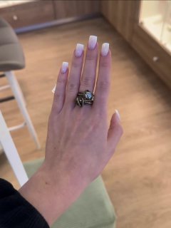 Крупный план руки с аккуратно накрашенными ногтями и двумя дизайнерскими кольцами из коллекции; интерьер магазина с деревянным полом на заднем плане.