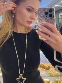 Селфи модели в зеркале с длинной цепочкой и серьгами Ciclon; витрина с украшениями Rosone видна позади, образ подчёркнут чёрным платьем.