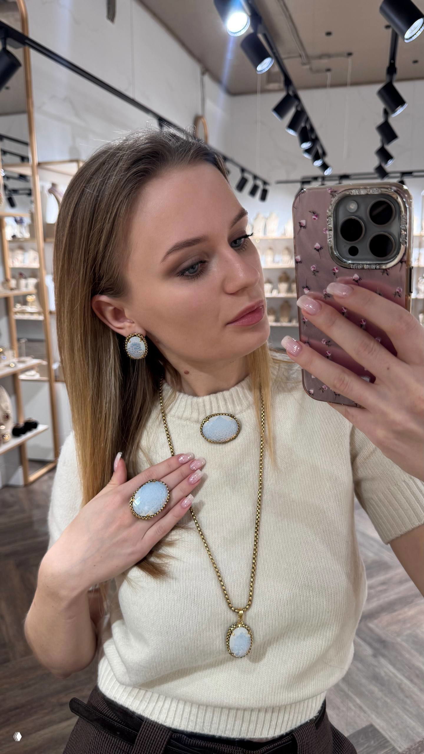 Модель в светлом свитере делает селфи в магазине, демонстрирует комплект с белыми вставками: серьги, кольцо и длинная цепочка с кулоном.