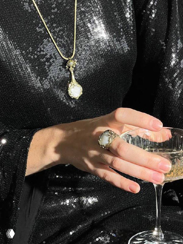Рука с крупным перламутровым кольцом и кулоном Fiore di Firenze на фоне чёрного блестящего платья и бокала шампанского, интимная деталь.