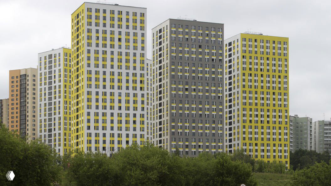 Фасады современных многоэтажных жилых домов с жёлто‑серой и серой отделкой на фоне пасмурного неба; внизу видны деревья и зелёная полоса.