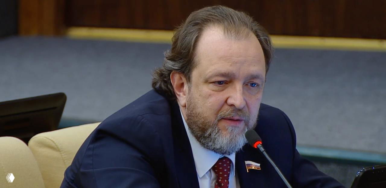 Депутат сидит в кресле и говорит в микрофон на фоне официального интерьера зала Госдумы; видны компьютерный монитор и трибуна.