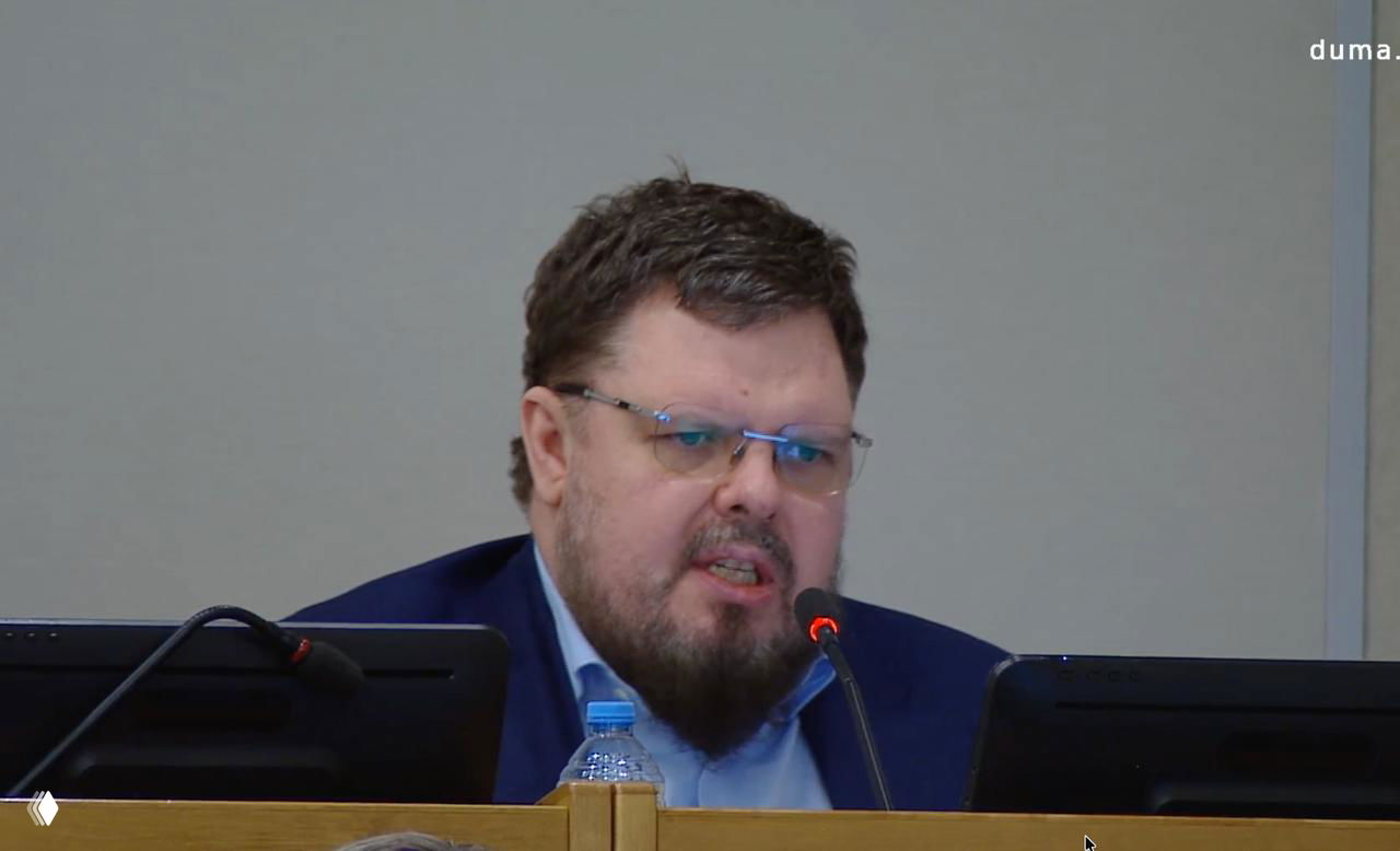 Крупный план выступающего депутата у микрофона в официальном зале заседаний; видны пульт, бутылка воды и часть трибуны в фоне.