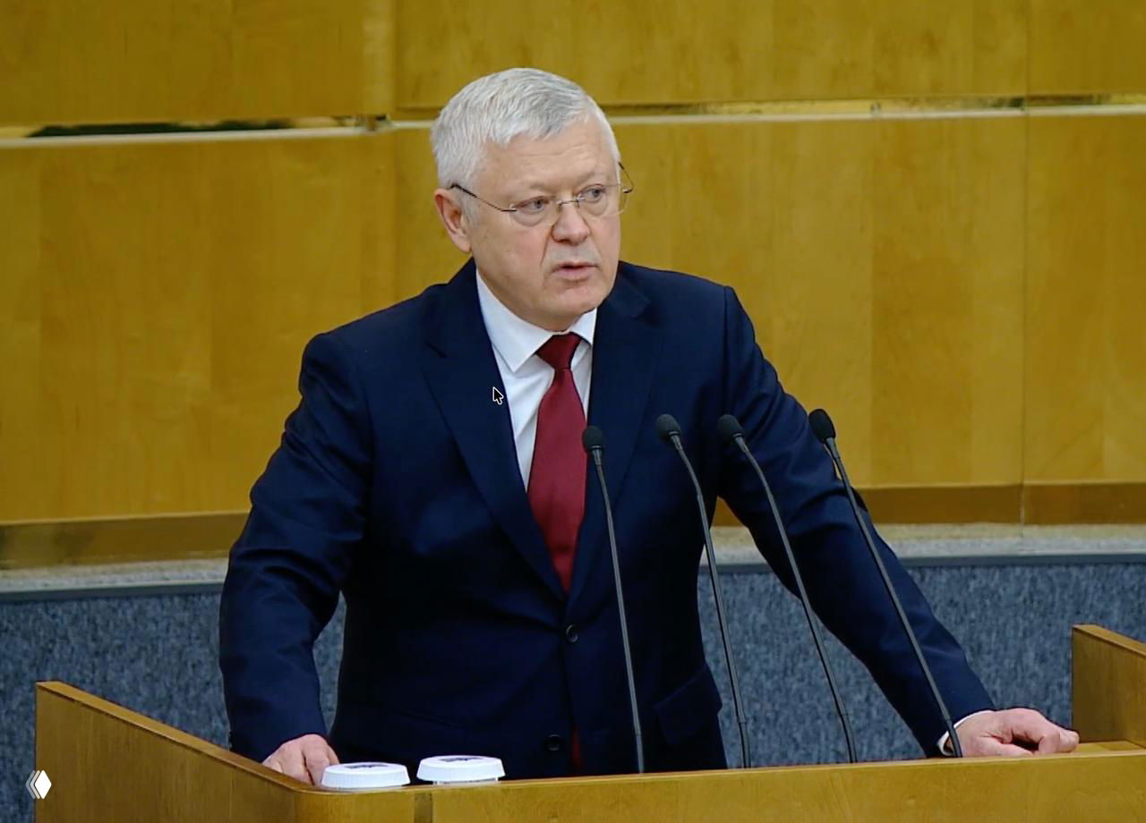 Депутат в деловом костюме выступает за трибуной в зале заседаний Госдумы; видны микрофоны, деревянные панели и официальный интерьер, заседание.