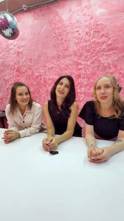 Три женщины сидят за столом на встрече агентства — улыбающиеся участницы или сотрудницы на розовом фоне, кадр по пояс.